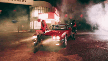 a woman sits on the hood of a red car in front of a building with a sign that says tomboy