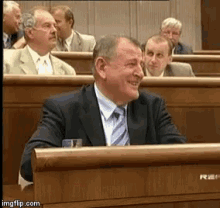 a man in a suit and tie is smiling while sitting at a podium in front of a group of men