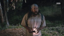 a man with a backpack is holding a flashlight in a dark forest with a hbo logo in the corner