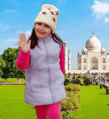 a little girl wearing a white hat and a gray vest is waving in front of the taj mahal