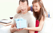 a woman covering a man 's eyes while he holds a gift