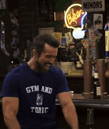 a man with a beard wearing a gym and tonic t-shirt is sitting at a bar .