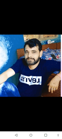 a man with a beard is sitting on a bed wearing a blue shirt that says ' river ' on the front .