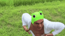 a man wearing a green frog hat and a white shirt is kneeling in the grass .