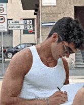 a man in a white tank top writes on a piece of paper in front of a sign that says s. marco pontaccio