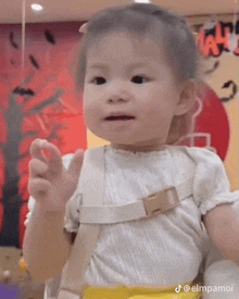 a baby girl wearing a white shirt and suspenders is sitting on a chair and making a funny face .