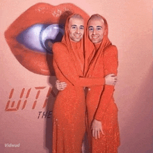 two men in orange dresses are hugging in front of a wall that says " with " on it