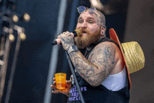 a man with a beard and tattoos is singing into a microphone while holding a cup of beer .