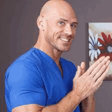 a bald man wearing a blue scrub top is clapping his hands in front of a painting of flowers .