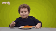 a little boy is sitting at a table with his hands in the air in front of a plate of food sponsored by onedio