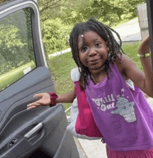 a little girl wearing a purple tank top that says chill out is standing next to a car door .