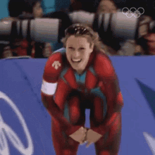 a woman in a red suit is smiling in front of a blue wall with the olympic rings on it