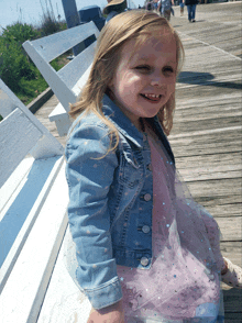 a little girl wearing a denim jacket and a pink dress smiles