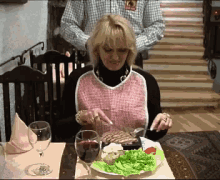 a woman wearing an apron is sitting at a table with a plate of food and a glass of wine