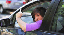 a man in a purple shirt is sticking his arm out of a car window and giving a middle finger .