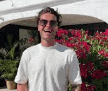 a man wearing sunglasses and a white t-shirt is standing in front of flowers and smiling .