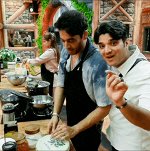 two men are preparing food in a kitchen and one of them is wearing an apron that says vegetables on it