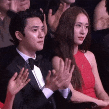 a man in a tuxedo and bow tie is sitting next to a woman in a red dress who is clapping .