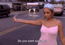 a woman in a pink tank top is standing on the side of the road holding up her hand and asking if she wants sausage .