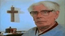 an elderly man wearing glasses is looking at the camera in front of a cross .