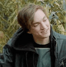 a young man in a leather jacket is smiling while standing in front of a bush .