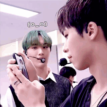 a man with green hair looks at another man holding a cellphone