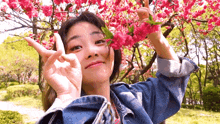 a woman in a denim jacket is making a peace sign in front of a tree with pink flowers .