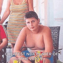 a shirtless boy is sitting on a chair with chips in his hand and a woman standing behind him .