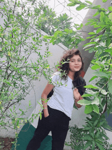 a woman in a white t-shirt and black pants is standing in front of a tree