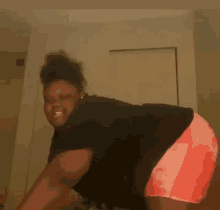 a woman in a black shirt and orange shorts is kneeling down in a room .