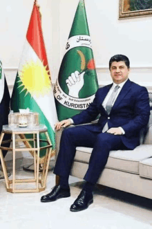 a man in a suit sits on a couch in front of flags with the word kurdistan on them