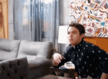 a man in a black shirt is sitting in a living room with a painting on the wall behind him