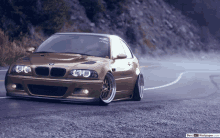 a brown bmw is driving down a road with a best hd wallpaper.com watermark