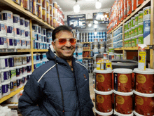 a man wearing sunglasses stands in front of a stack of buckets of paint