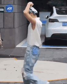 a man in a white tank top and blue jeans is standing in front of a white tesla model s .