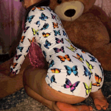 a woman wearing a white bodysuit with butterflies on it is kneeling next to a teddy bear