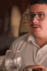 a man with glasses and a mustache is sitting at a table eating a hamburger