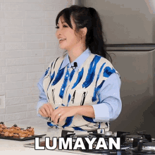 a woman in a blue and white vest is smiling in front of a stove with the word lumayan written on it