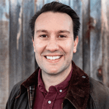 a man wearing a brown jacket and a red shirt smiles in front of a wooden wall