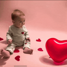 a baby is sitting next to a large red heart shaped balloon