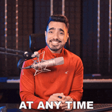 a man in a red shirt sitting in front of a microphone with the words at any time below him