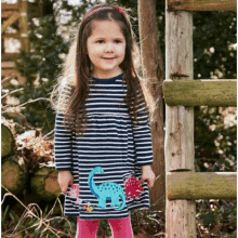 a little girl wearing a striped dress with a dinosaur on it is standing in front of a wooden fence .