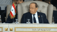 a man in a suit and tie sits at a table with a small lebanon flag in front of him