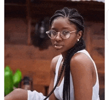 a woman with braids and glasses is sitting in front of a wooden building and looking at the camera .