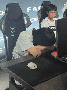 a man in a white shirt with the word sho on it sits at a desk with his head down