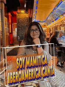 a woman sits at a table in front of a sign that says soy mamutica iberoamerica canta