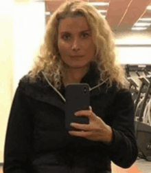 a woman in a black jacket is taking a selfie with her cell phone in a gym .