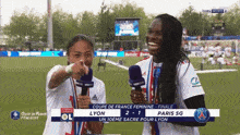 two women holding microphones on a soccer field with a scoreboard in the background that says lyon 2-1 paris sg