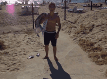 a shirtless young man standing on a beach holding a towel