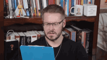 a man wearing glasses looks at a clipboard in front of a bookshelf with a mug that says contains one ordinary coat